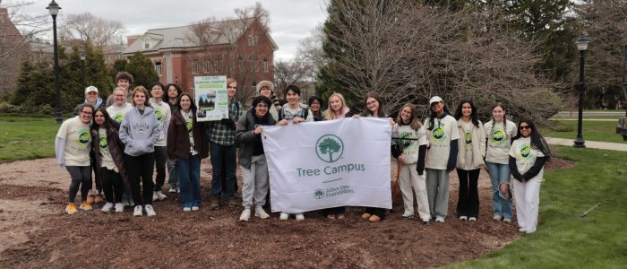 A group of students pose in a mulch ring in front of the new Class of 2028 tree and pose with the Tree Campus USA banner